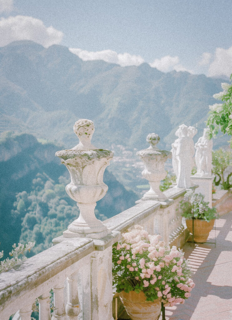 Full-service wedding planning for busy professionals 1 Ravello, Italy in the style of film photography with a vintage vibe. The wedding scene is set on an outdoor terrace overlooking mountains and sea, featuring white stone sculptures of classical figures along one side of the balcony.